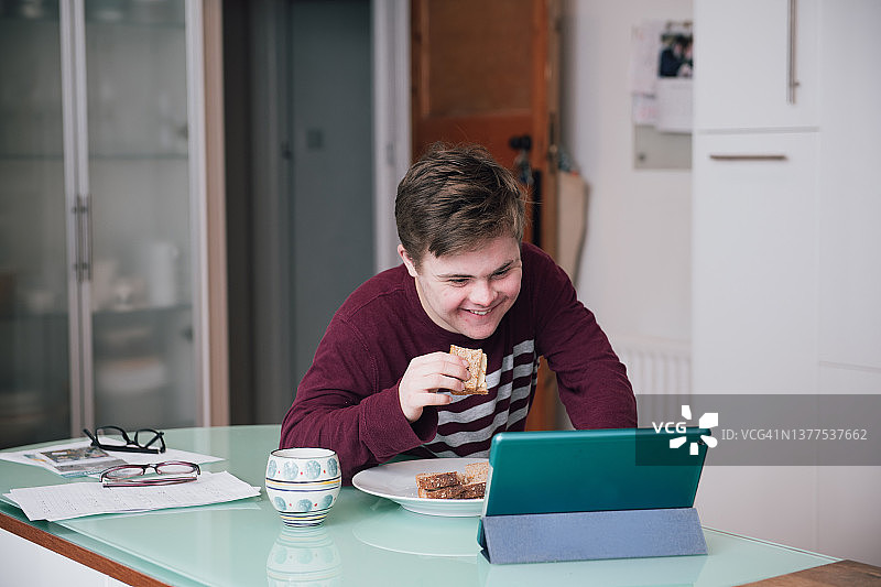 患有唐氏综合征的青少年男孩边吃三明治边在平板电脑上看电影图片素材