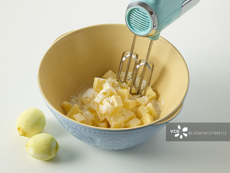 用蓝色电动搅拌器在大号蓝色搅拌碗中将黄油、细砂糖和柠檬皮混合，制作裸蛋糕，旁边放磨碎的柠檬图片素材