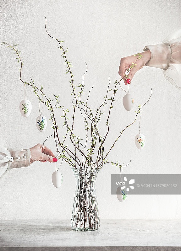 涂着红色指甲油的双手装饰着玻璃花瓶中的春天树枝，灰色桌子上有白色复活节彩蛋图片素材