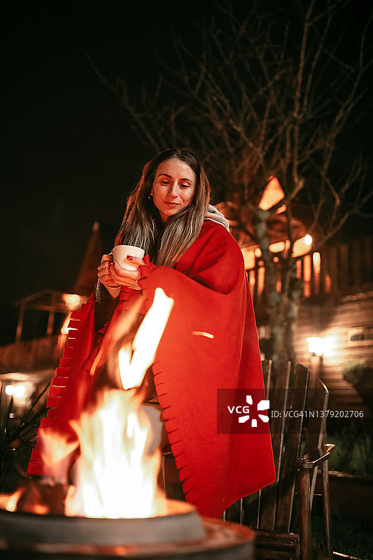 快乐的女人披着红色披肩在燃烧的篝火旁喝咖啡，女性生活方式图片素材