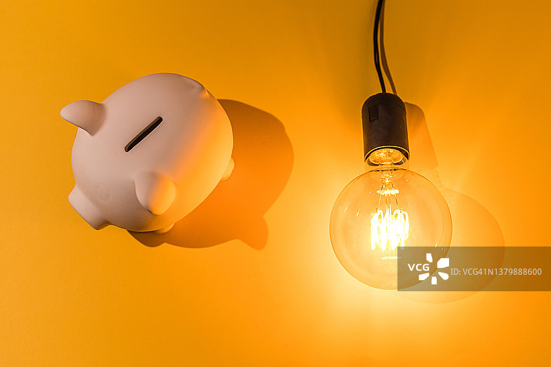 粉色背景上点亮的灯泡旁的储蓄罐，电力价格、危机、金钱、储蓄和电力能源概念图片素材