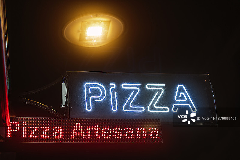 蓝色、红色和橙色霓虹灯比萨店招牌夜景。西班牙语文字：手工比萨。黑暗的复制空间图片素材