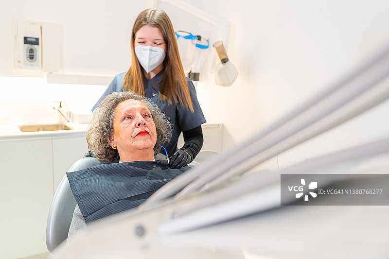 牙科诊所内，助手戴着口罩为老年女顾客戴围嘴，协助其进行牙科治疗图片素材