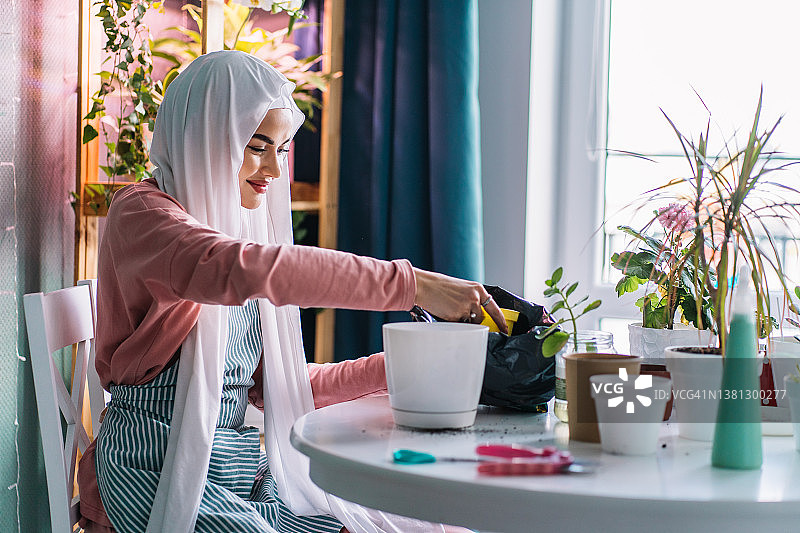 微笑着安静的穆斯林妇女在白色面纱面纱盖她的头，与化妆，坐在和园艺室内。再播绿党。图片素材