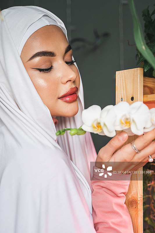 面带微笑、有魅力的穆斯林妇女，戴着白色头巾，手持并嗅着新鲜的兰花，准备过生日节日图片素材
