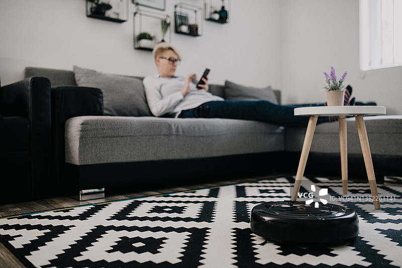 女士在沙发上休息时，机器人真空吸尘器正在清洁房间。女士使用遥控器控制吸尘器。图片素材
