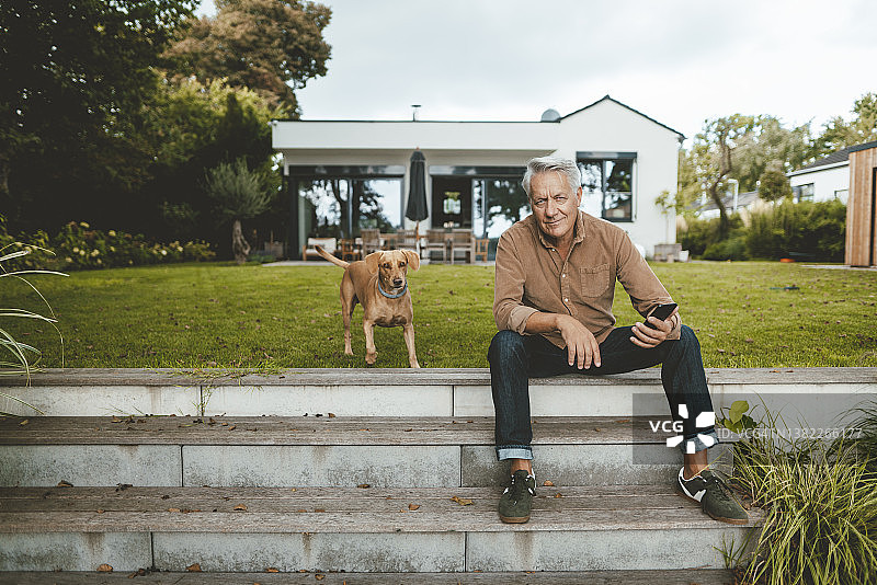 坐在后院台阶上，与狗在一起玩智能手机的年长男人图片素材