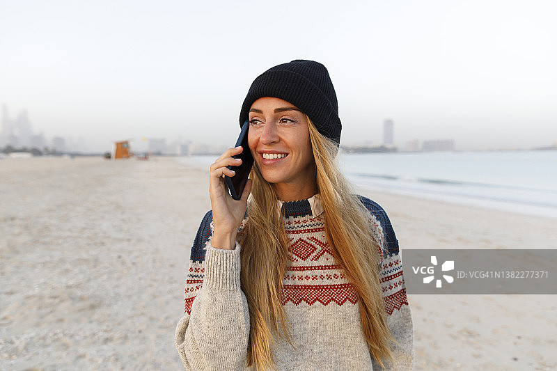微笑女人在海滩戴着黑色针织帽用智能手机通话图片素材