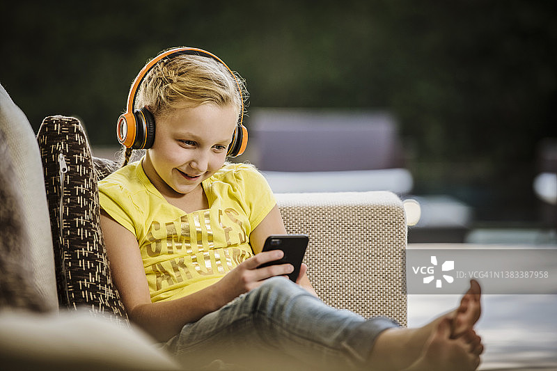 戴着耳机和智能手机坐在沙发上的微笑女孩（10-11岁）图片素材