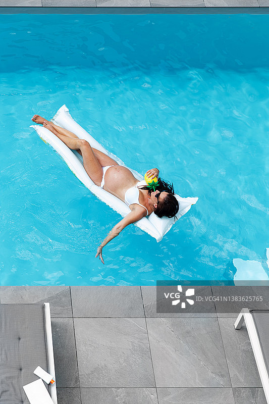 孕妇漂浮在游泳池的充气垫上，手拿热带饮品图片素材