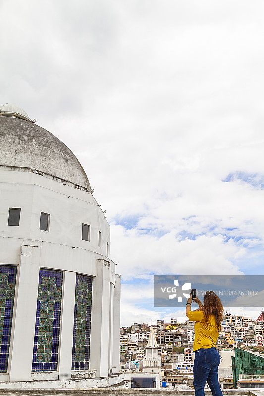 中年拉丁裔女游客在厄瓜多尔安巴托大教堂顶部旅行、放松、享受并拍照；背景是晴朗多云的城市景观图片素材