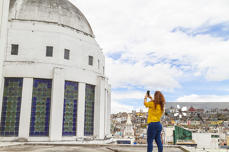 中年拉丁裔女游客在厄瓜多尔安巴托大教堂顶部旅行、放松、享受并拍照；背景是晴朗多云的城市景观图片素材