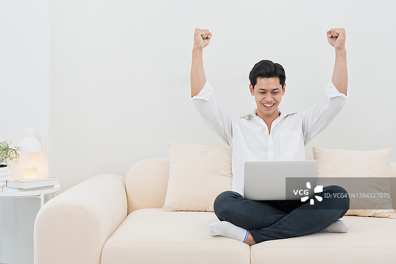 微笑的亚洲男人在沙发上使用笔记本电脑，成功和快乐情绪概念图片素材