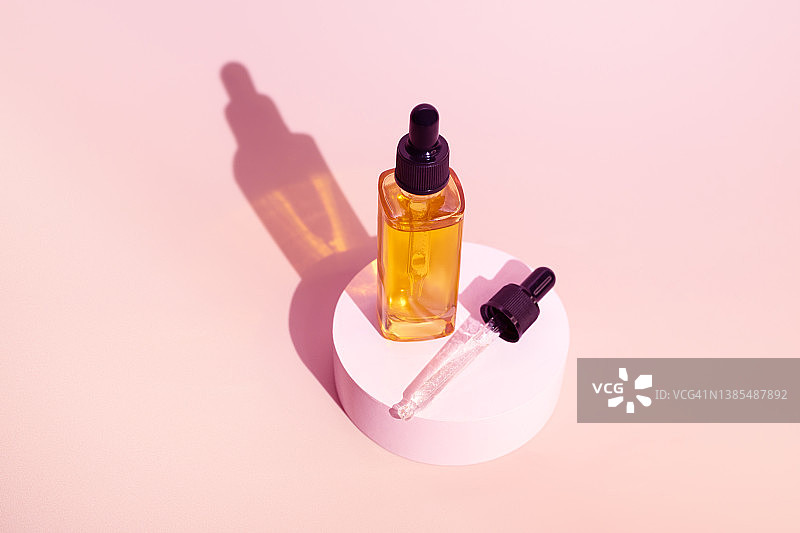 粉色背景圆形展台上带滴管的精华玻璃瓶：天然有机水疗化妆品概念，正面视角图片素材