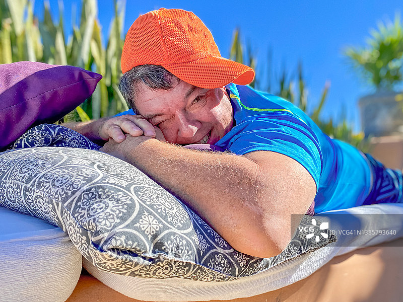 微笑的中年男人戴着棒球帽，在阳光下枕着枕头放松图片素材