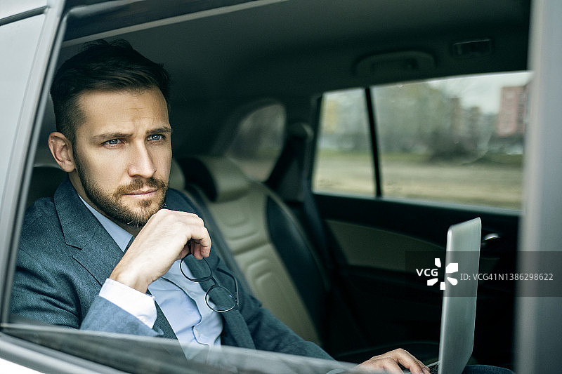 商人坐在豪华轿车后座，使用笔记本电脑，透过窗户向外看图片素材