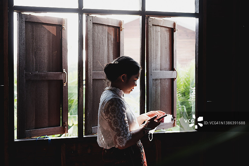 身着传统服饰的泰国女性在窗边手持智能手机和面具图片素材