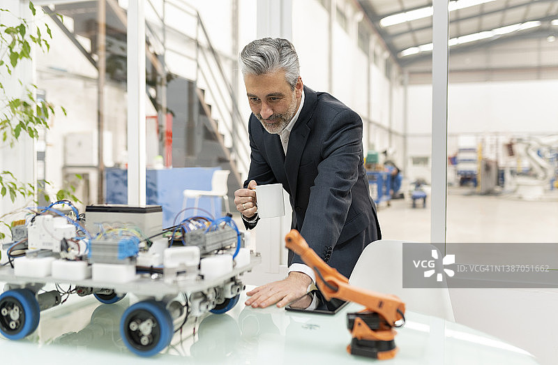 手持咖啡杯查看办公桌上的机器人汽车模型的商人图片素材