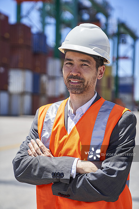 亚裔美国男工程师戴着安全帽和反光背心站在集装箱堆前图片素材