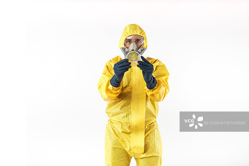 穿着黄色核防护服、戴着面罩和防护眼镜的技术员拿着一个带有植物样本的培养皿，在白色背景上。核能与疫情的概念。图片素材