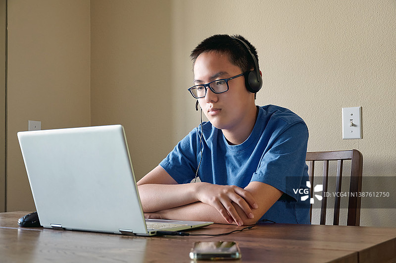戴着耳机在桌边使用笔记本电脑的东亚裔男孩图片素材
