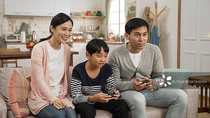 亚洲父亲和儿子在家中客厅一起用手柄玩电子游戏，母亲加入并欢呼图片素材