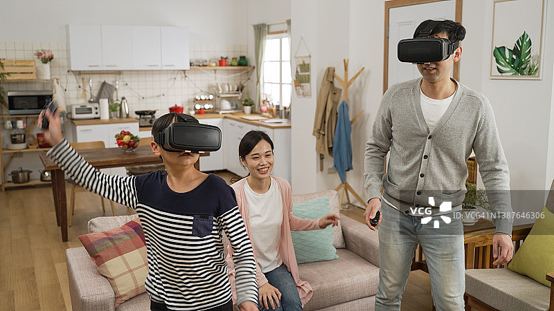 幸福的亚洲家庭在客厅里玩耍，母亲看着儿子和父亲使用VR头显玩游戏。图片素材
