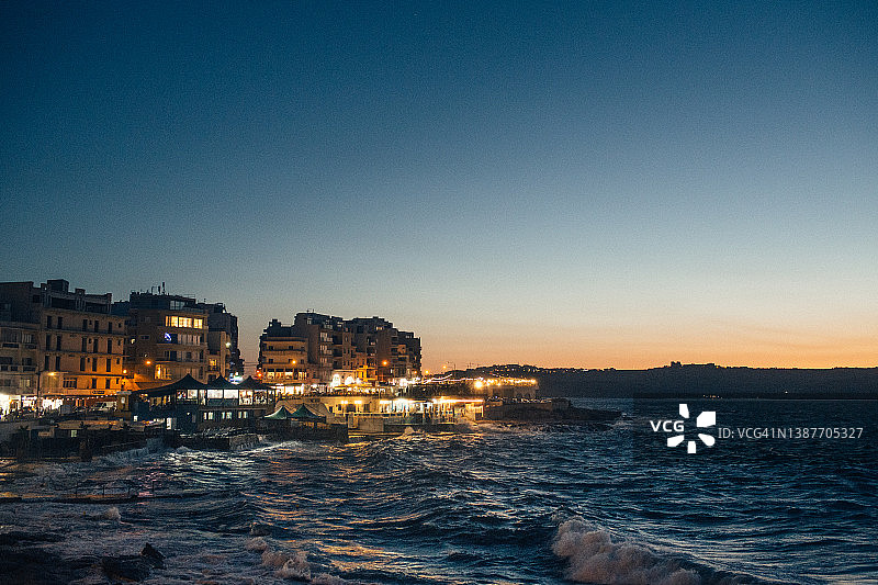马耳他布吉巴的夜间沿海景色，餐厅灯光和远处日落，海浪滔天图片素材
