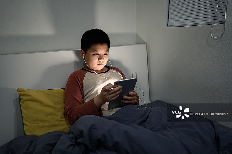 英俊的亚洲男孩坐在床上听音乐，看着平板电脑图片素材