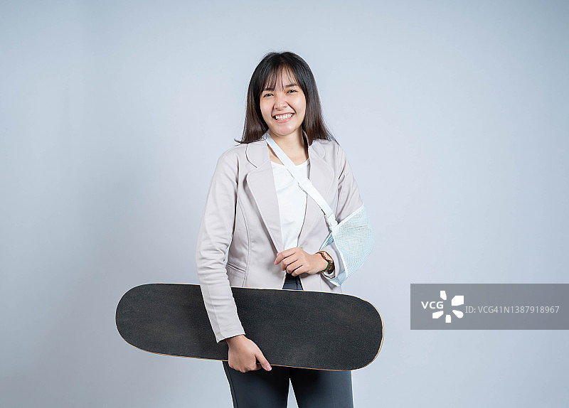 手臂骨折后微笑的女性，佩戴手臂夹板接受治疗，玩滑板运动受伤，快乐的亚洲女性用吊带支撑手部，灰色背景图片素材