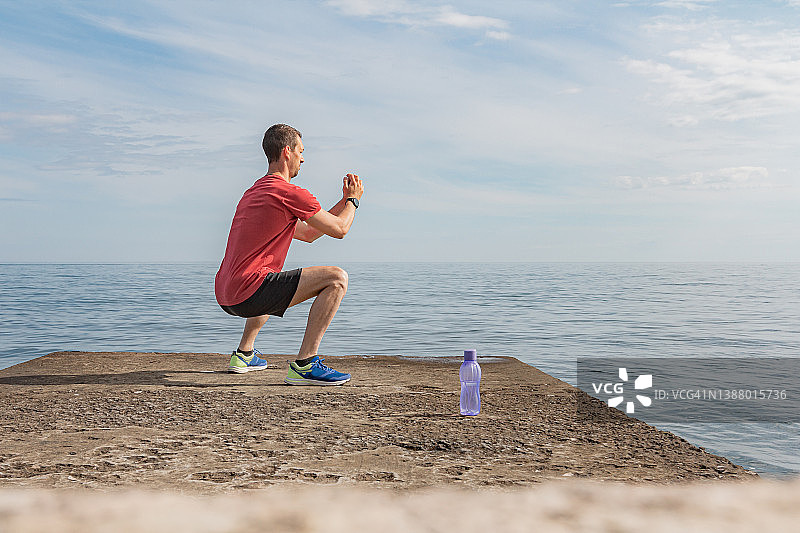 身材健壮的男人穿着红色T恤在海边锻炼并做下蹲图片素材