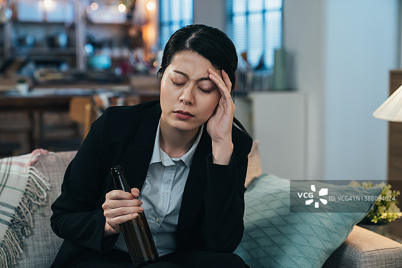 头痛难忍的亚洲醉酒女商人。手持啤酒瓶的韩国女性正揉着太阳穴，表情难受。图片素材