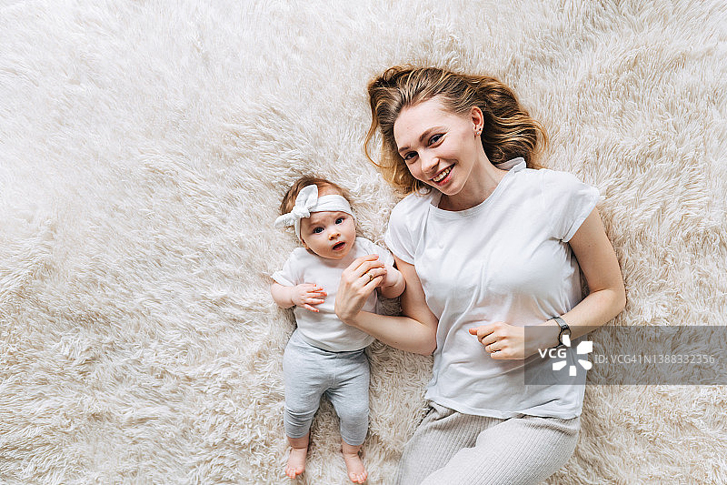 一位年轻微笑的温柔金发长发母亲与女儿一起躺在毛皮毯子上，看着镜头图片素材