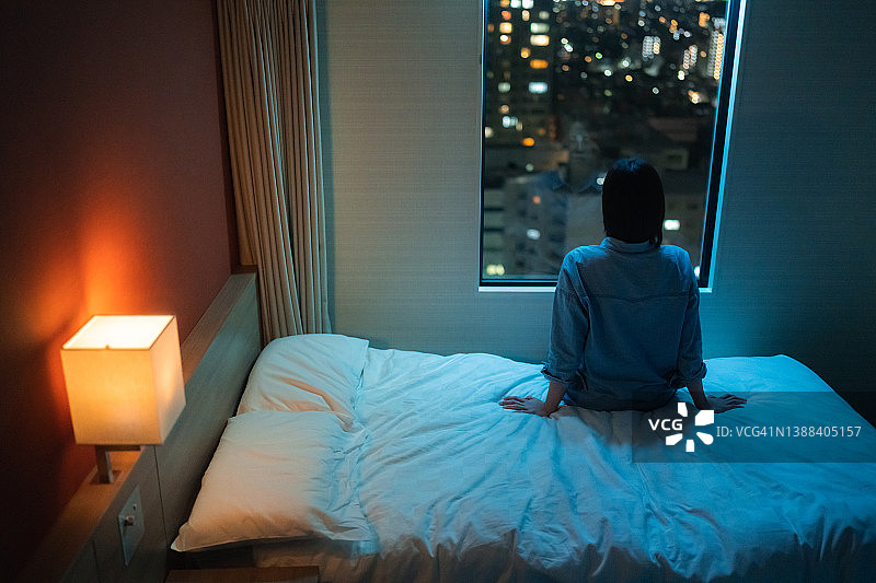 夜晚，女子独自坐在房间的床上，望着窗外的背影图片素材