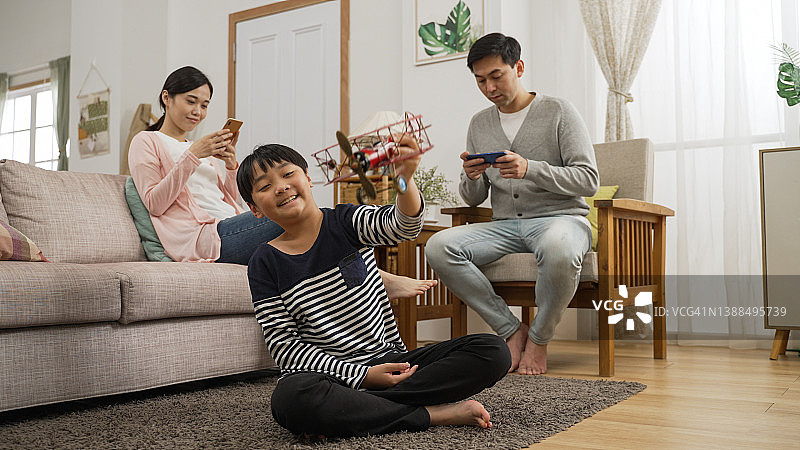 快乐的学龄儿童玩着玩具飞机，梦想成为飞行员。父母在家玩手机图片素材