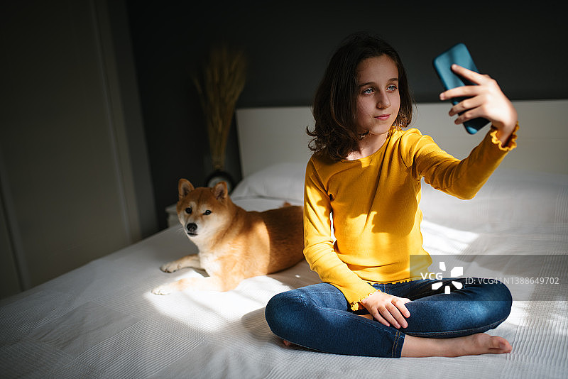 坐在床上用手机和她的狗自拍的年轻女孩图片素材