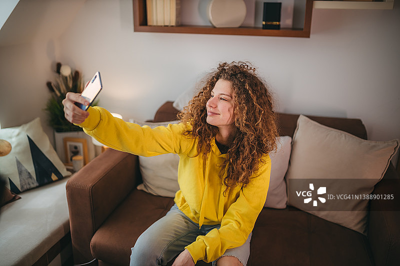 年轻卷发女人在会议视频中使用手机，在沙发上放松图片素材