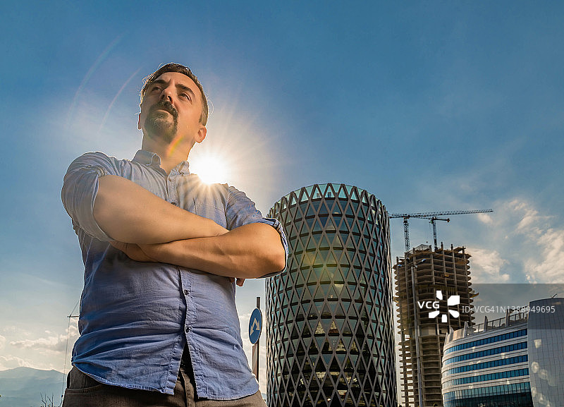 在现代商业建筑和建筑工地前，交叉双臂站立的年轻男人图片素材
