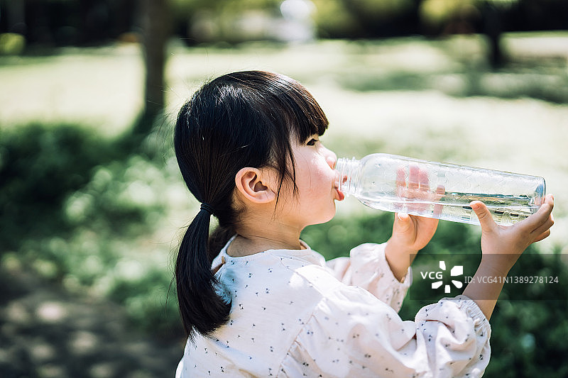 阳光明媚的日子里，亚洲小女孩在公园户外用可重复使用的塑料瓶喝水的侧面照。零浪费和可持续生活方式概念图片素材