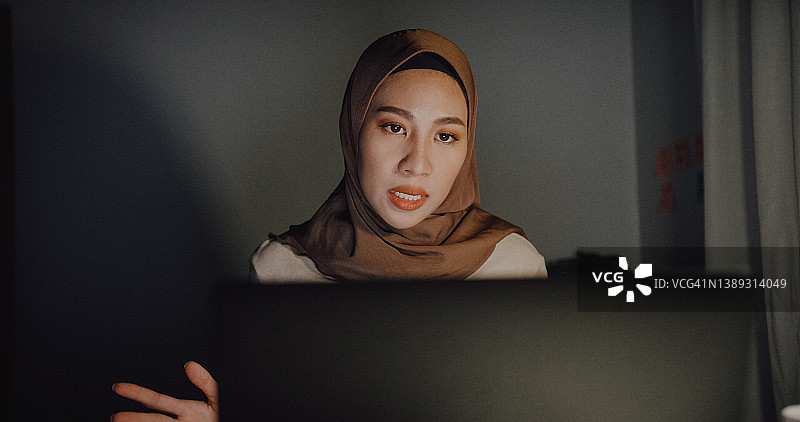 年轻的亚洲穆斯林女商人戴着头巾在家中晚上坐在桌旁通过笔记本电脑进行视频通话图片素材