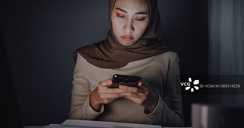 年轻的亚洲穆斯林女商人戴着头巾，晚上在家里的办公桌上用笔记本电脑工作和使用手机图片素材