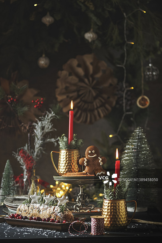 圣诞静物背景：传统圣诞蛋糕、带红色蜡烛的铜杯、姜饼人、圣诞树和悬挂的纸星星，深色桌子上有散景灯光图片素材