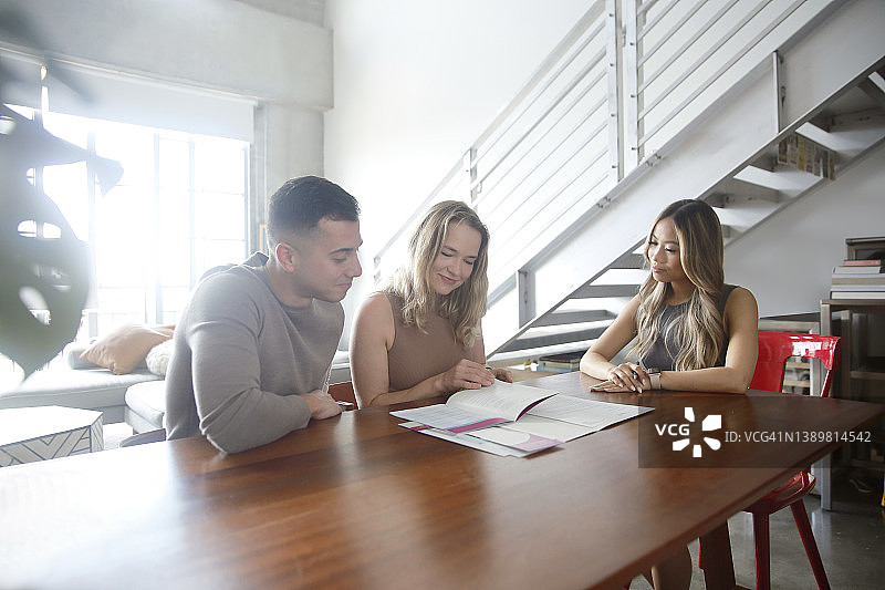 一对千禧一代的白人夫妇在光线明亮的阁楼公寓里与一位亚洲女房地产经纪人会面，签署文件，身穿时尚休闲服装图片素材