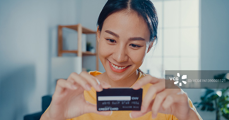年轻亚裔女子手持信用卡，使用笔记本电脑在家中客厅的办公桌旁进行在线支付图片素材