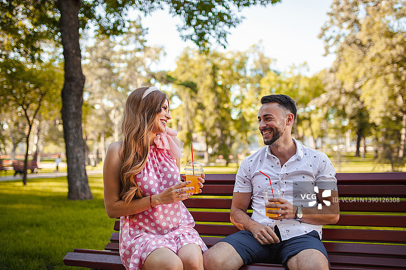 在一个温暖的夏日，年轻夫妇在公园长椅上放松身心，喝着橙汁图片素材