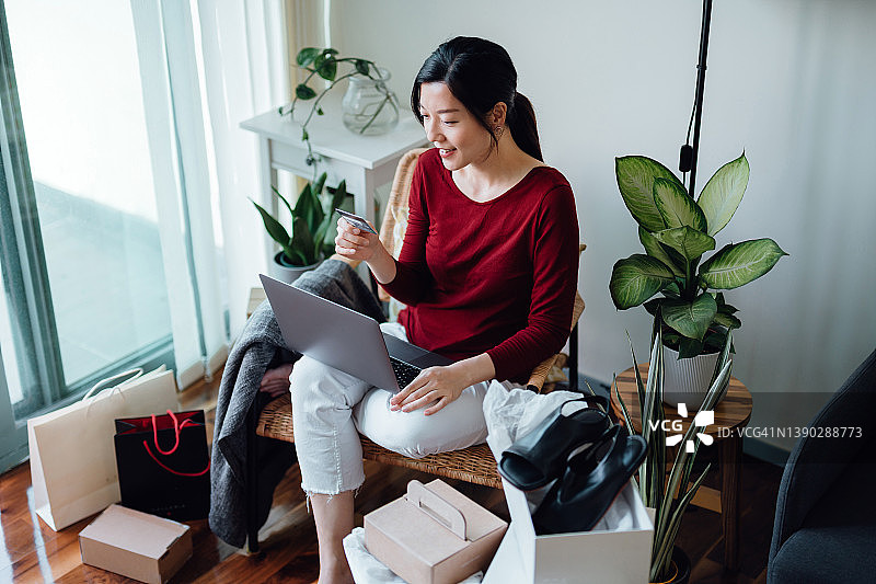 面带微笑的亚洲年轻女子在家用笔记本电脑在线购物，用信用卡在线支付，旁边放着购物袋和快递包裹。生活方式与科技。在线购物与电子商务。非接触式支付概念图片素材