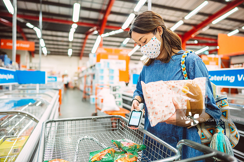 年轻亚洲女性戴防护口罩在超市使用手持条形码扫描器购物图片素材