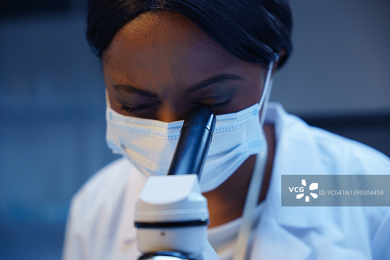 戴防护口罩的年轻非洲裔美国医生在模糊背景下观察显微镜图片素材