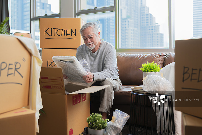 一位年长的亚洲男士坐在地板上，准备搬到新家，将物品打包到盒子里，并在客厅里用胶带封住盒子，搬家创意概念图片素材