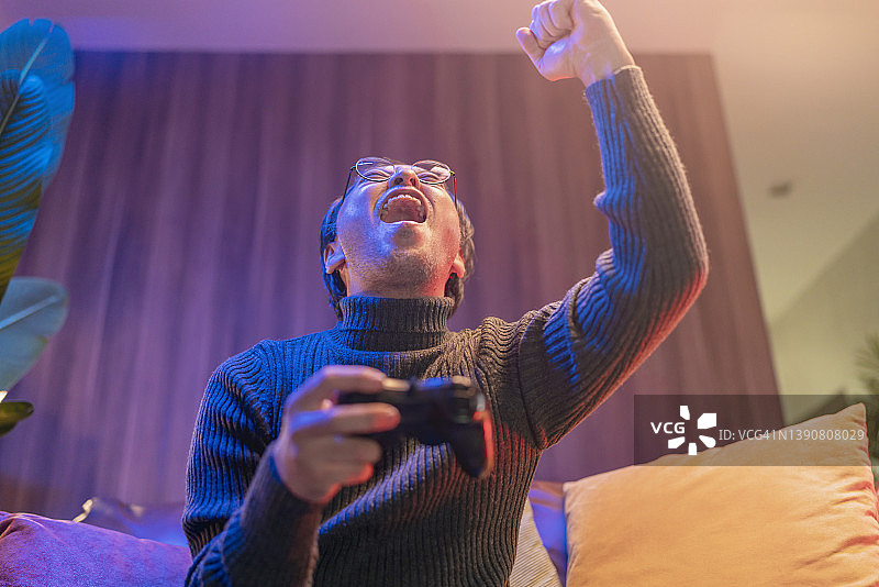 兴奋的亚洲日本中年男子独自在家中黑暗中玩电子游戏，坐在沙发上的游戏控制器，亚洲男性反应享受他在游戏中的胜利，举起手在空中欢呼，并在夜间致以获胜者问候图片素材